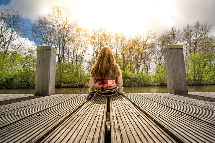 woman sitting down on brown wooden dock facing lake waters