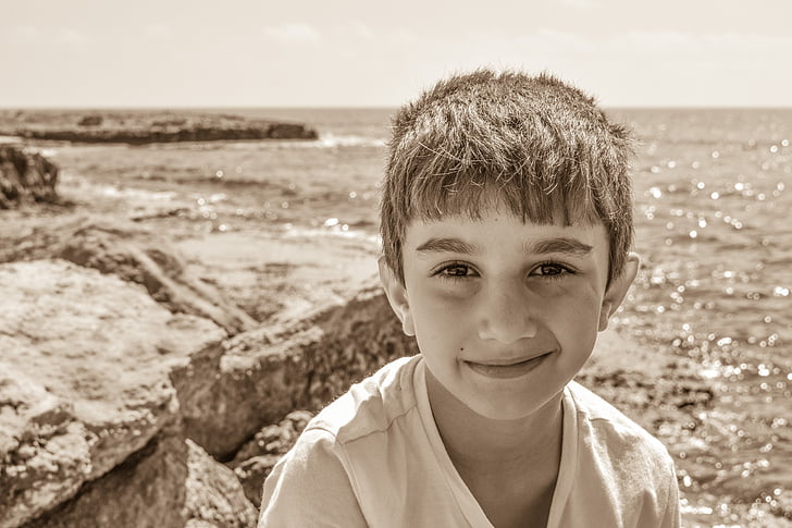 sepia photography of boy standing on cliff mountain fronting body of water