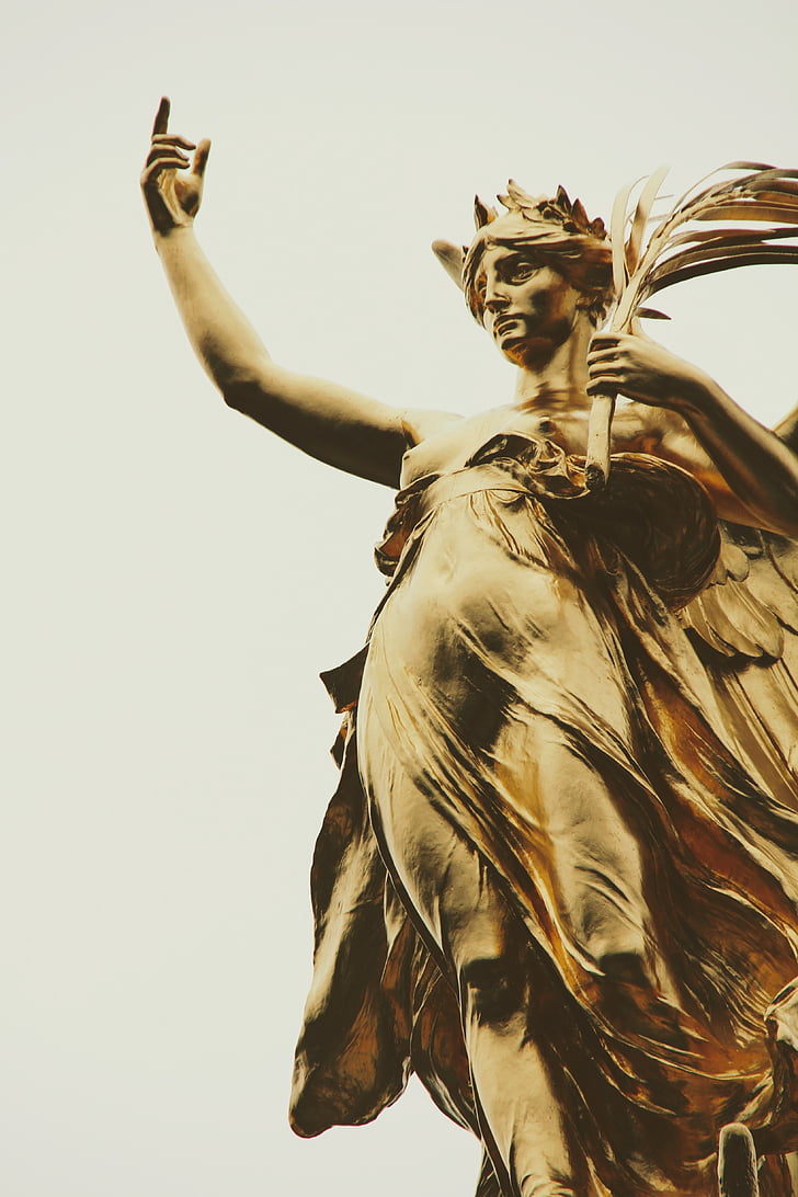 woman raising her right hand while holding branch with leaves on her left hand brass-colored statue low-angle photo