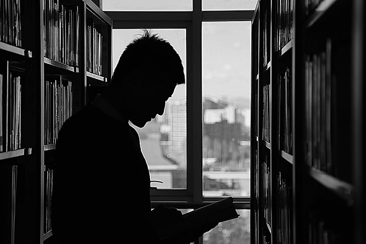 silhouette of man reading book in library near window panes