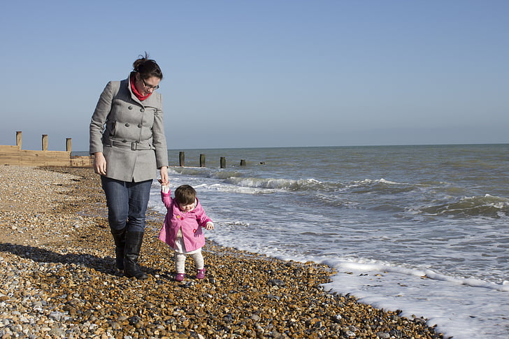 woman in grey double-breasted coat and toddler in pink jacket walking hand-in-hand at beach at daytime