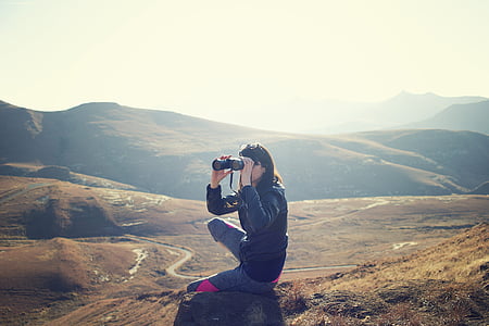 woman sitting on rock while using binoculars during daytime