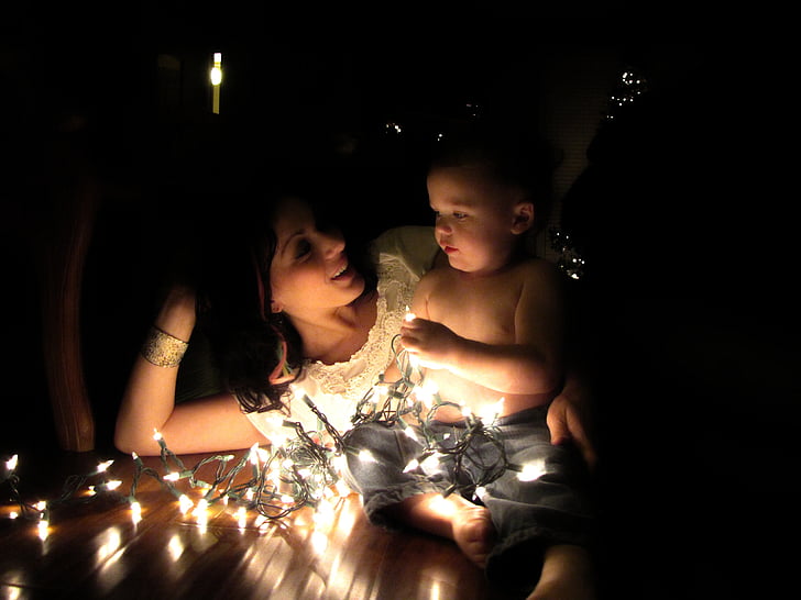 photo of woman laying on brown wooden floor with topless baby holding stringed lights