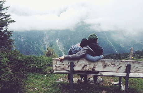 two person wearing jackets sitting on bench staring at mountain covered with trees and fog