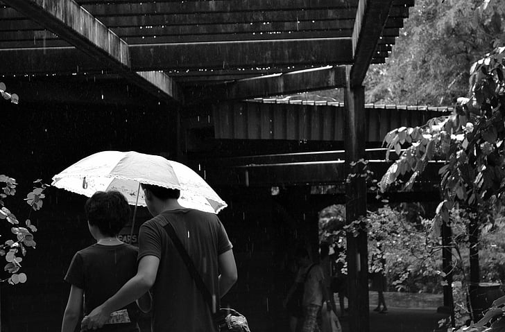 grayscale photo of man and woman under umbrella near brown wooden building
