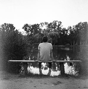 grayscale photography of man in shirt sit on bench in front of lake