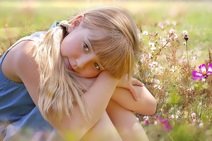 selective focus of girl wearing blue dress sitting on ground surrounded by flowers