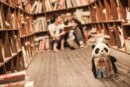 selective focus photography of panda plush toy holding a book in front of two person sitting on bookcase