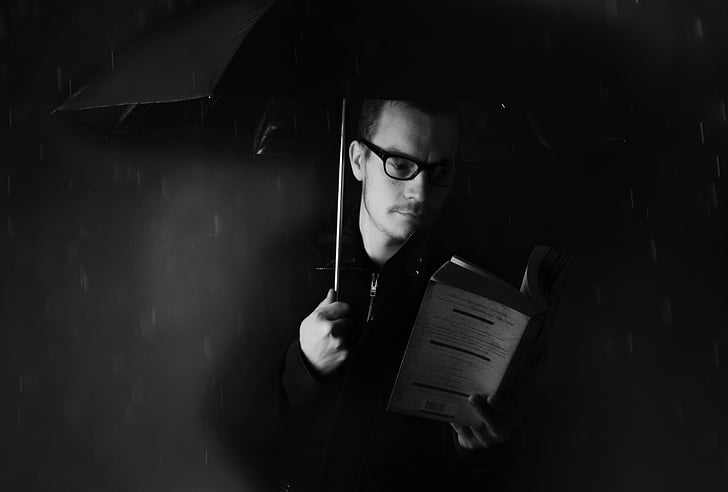 grayscale photograph of man reading book under umbrella during rain