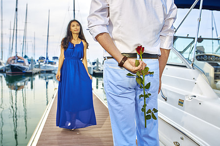 woman in blue sleeveless dress and man in white dress shirt and blue jeans holding red rose behind his back on pier bridge during daytime