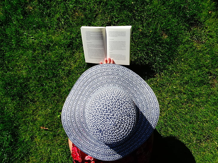 photograph of person wearing sun hat reading book on field of grass