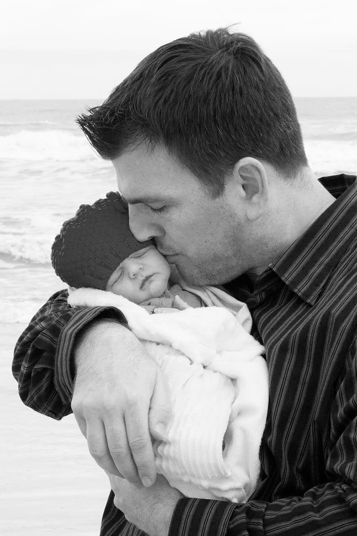 grayscale photo of man kissing baby wrapped in towel on seashore