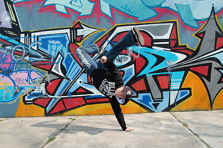 man wears black hoodie doing one hand stand near multicolored wall graffiti during daytime