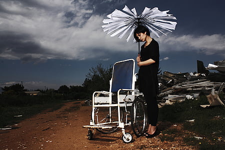 woman standing beside empty wheelchair holding broken umbrella