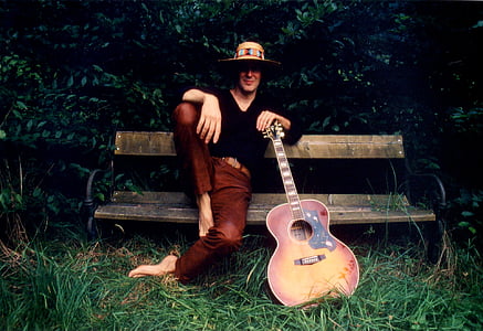 man in black long-sleeved shirt and brown pants sitting on brown wooden bench with guitar