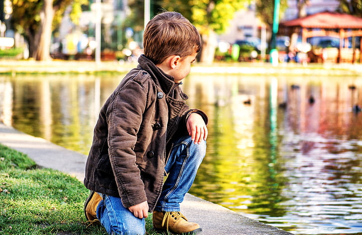 boy kneeling on green grass beside body of water at daytime