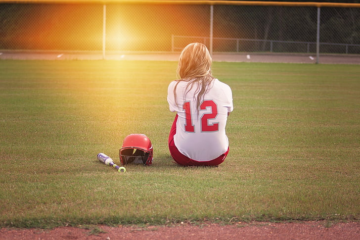 woman in white and red 12 jersey sitting on the field beside baseball bat and batting hat