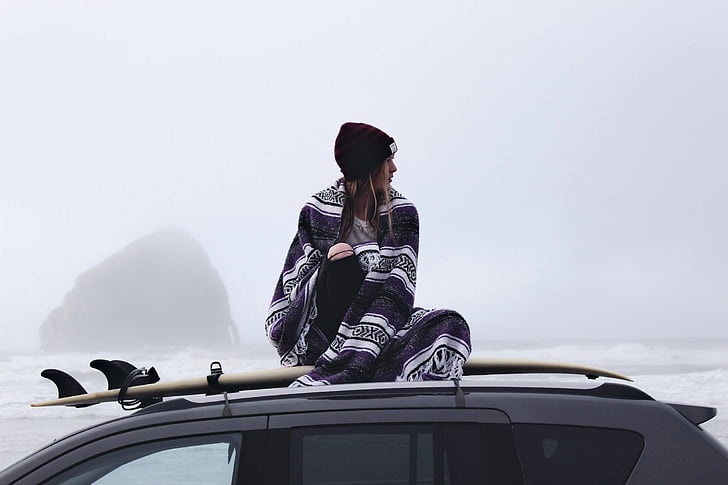 woman sitting on top of gray SUV with surfboard