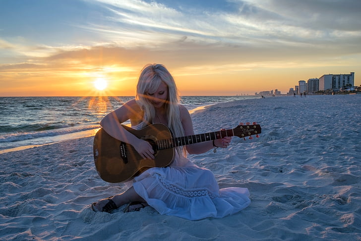 woman in white dress sitting on white sand while playing a guitar