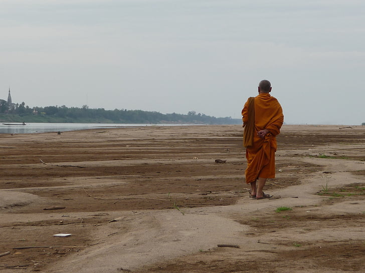 monk walking on soil beside water and forest