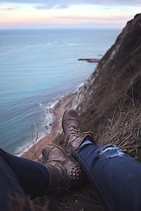 selective focus photography of person wearing pair of gray leather side-zipped combat boots sitting on edge of mountain by the sea
