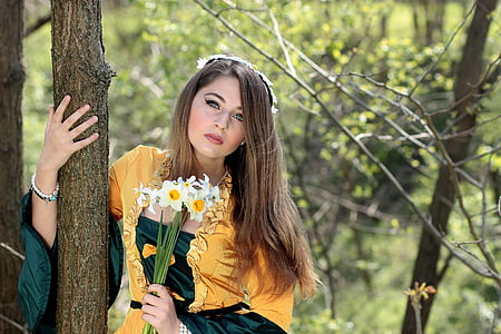 woman holding white petaled flowers standing beside brown tree trunk at daytime