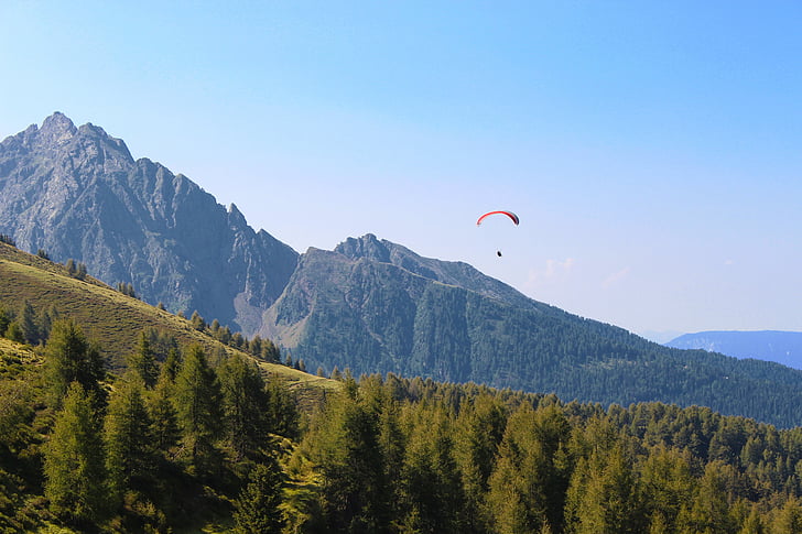 person parachuting down hillside with pine trees under blue sky during daytime