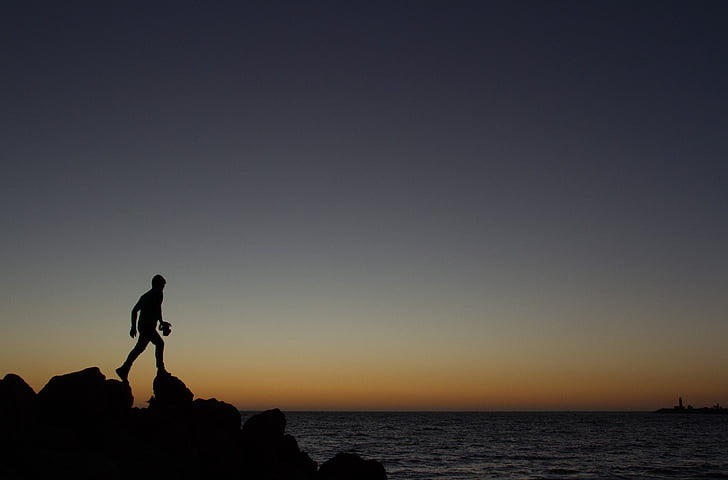 silhouette of person walking on rock at the edge of ocean during golden hour