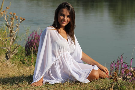 smiling woman wearing white v-neck flare-sleeved cover-ups sitting on green grass beside body of water during daytime