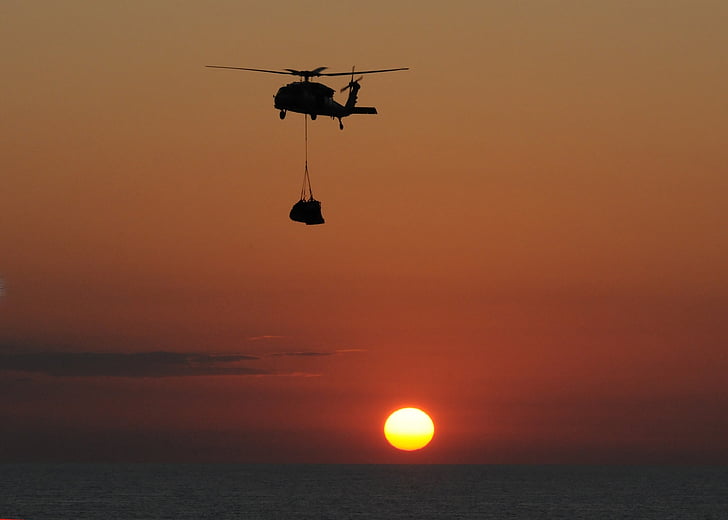 helicopter carrying cargo over body of water during golden hour