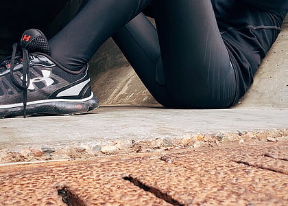 person wearing gray Under Armour running shoes laying on floor while leaning on concrete wall