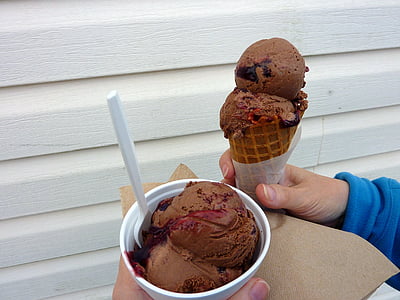 person holding chocolate flavor ice cream on cone and cup besides white wooden wall