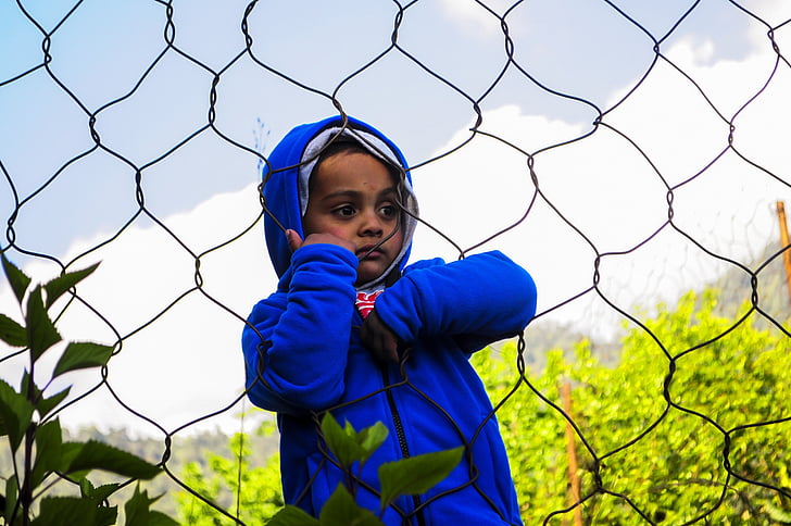boy wearing blue hoodie holding on link fence