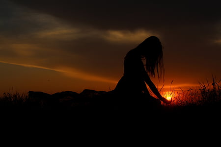 silhouette photography of woman sitting on grass under grey clouds orange skies sunset