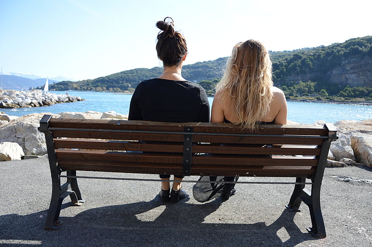 two women sitting on bench near body of water and mountains during daytime