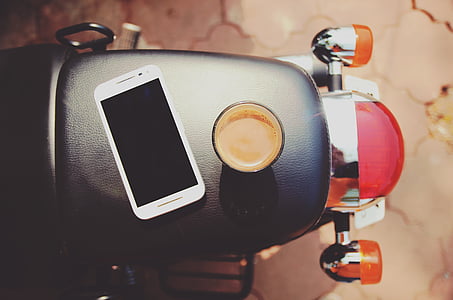 white Android smartphone beside black mug on top of black motorcycle seat