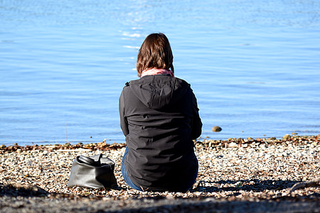 woman sitting on gray sand seashore wearing black hooded jacket