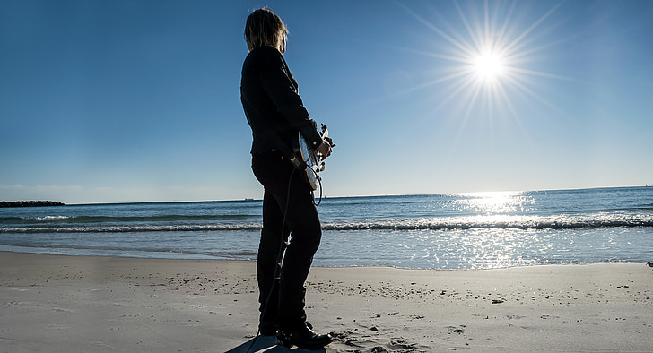 person playing guitar standing on shoreline facing on ocean