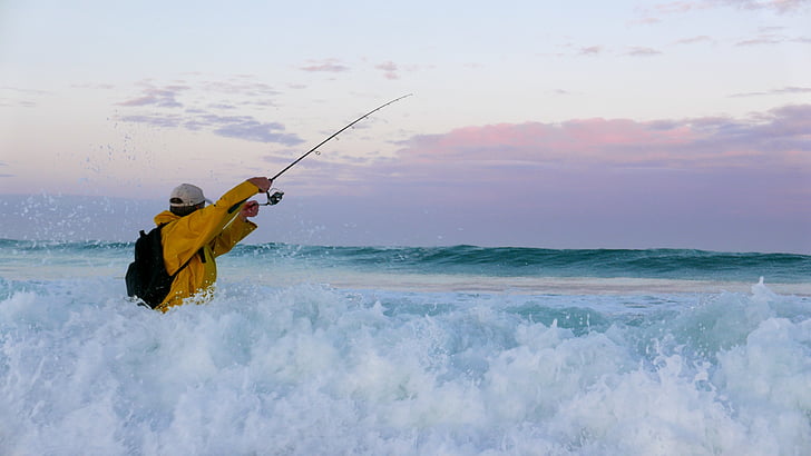 Royalty-Free photo: Man wearing orange windbreaker jacket holding telescopic  fishing rod on sea water