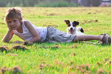 boy wearing white tank top lying on green grass beside short-coated white and brown dog during daytime