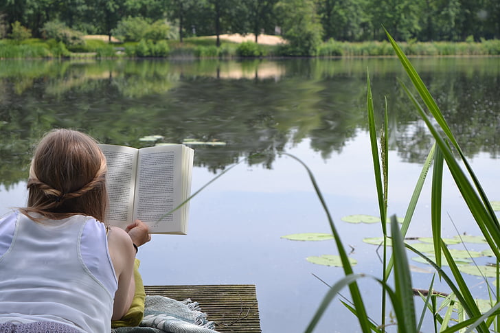 woman reading book on brown dock near lake