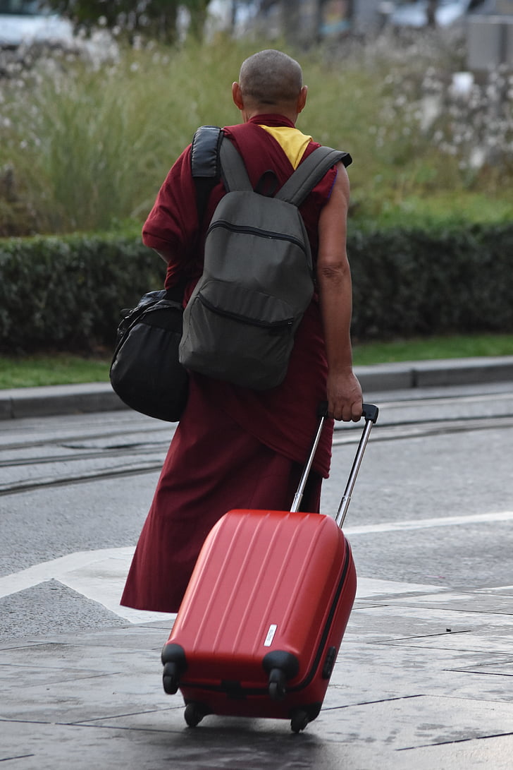monk carrying bag while holding red luggage walking on road