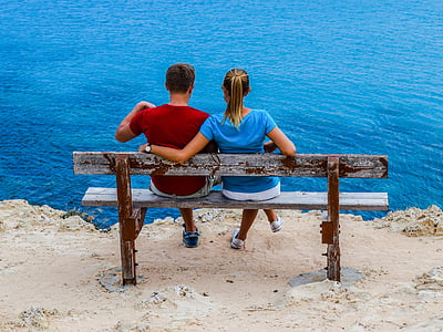 couple sitting on beige wooden bench on cliff by the water during daytime