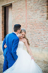 woman wearing white bridal gown beside man wearing blue suit jacket and pants near brown brick wall