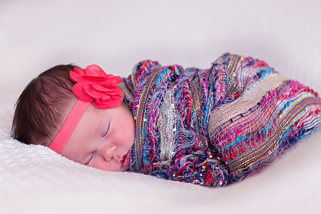 baby sleeping wearing multicolored blanket and red flower headband in closeup photography