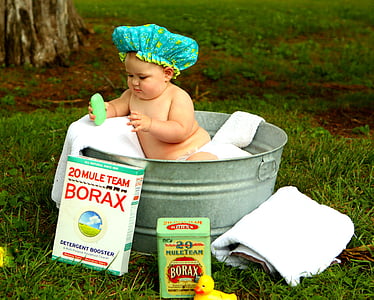 baby wearing green and yellow shower net in gray metal basin holding soap beside Borax boxes