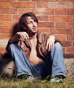 man wearing brown leather jacket and blue jeans sitting on grass near brick wall