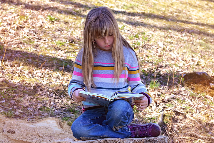 girl reading book sitting on green grass at daytime