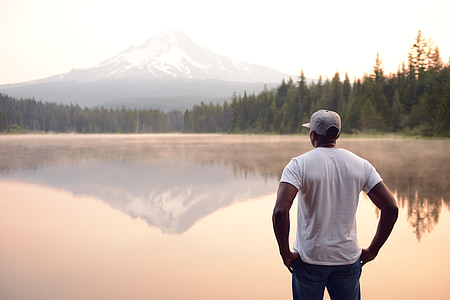 man in white shirt and blue jeans standing near lake and body of mountain during daytime