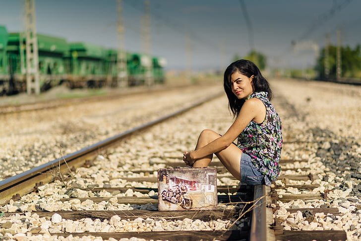 woman wearing black and multicolored sleeveless top and blue denim shorts outfit sitting on train railways under blue sky during daytime
